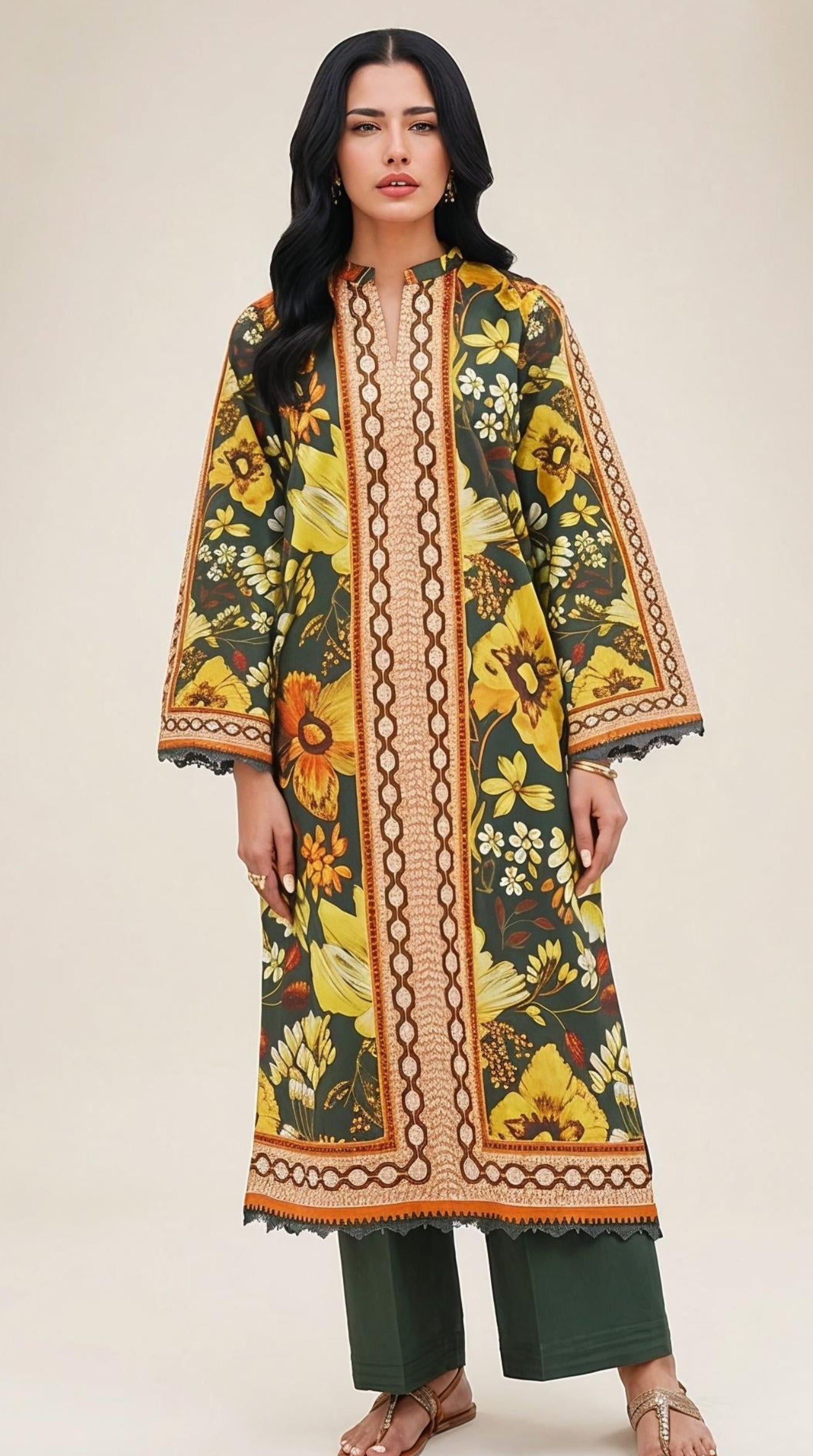 A woman modeling a long-sleeved, knee-length kaftan-style tunic with a vibrant yellow, green, and orange floral print and a central beige geometric border, paired with dark green wide-leg pants and brown sandals, set against a minimalist beige studio backdrop.