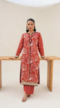 A woman models a rust-red three-piece kurta set with white floral embroidery and black trim, standing behind a light wood table in a minimalist studio setting.